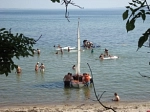 Das Bild wurde im Zeltlager aufgenommen, zu sehen ist der Blick vom Lagerplatz auf die Ostsee. Ein Segelboot liegt im Wasser, Kinder spielen auf der Badeinsel, andere schwimmen oder spielen auf Surfbrettern.